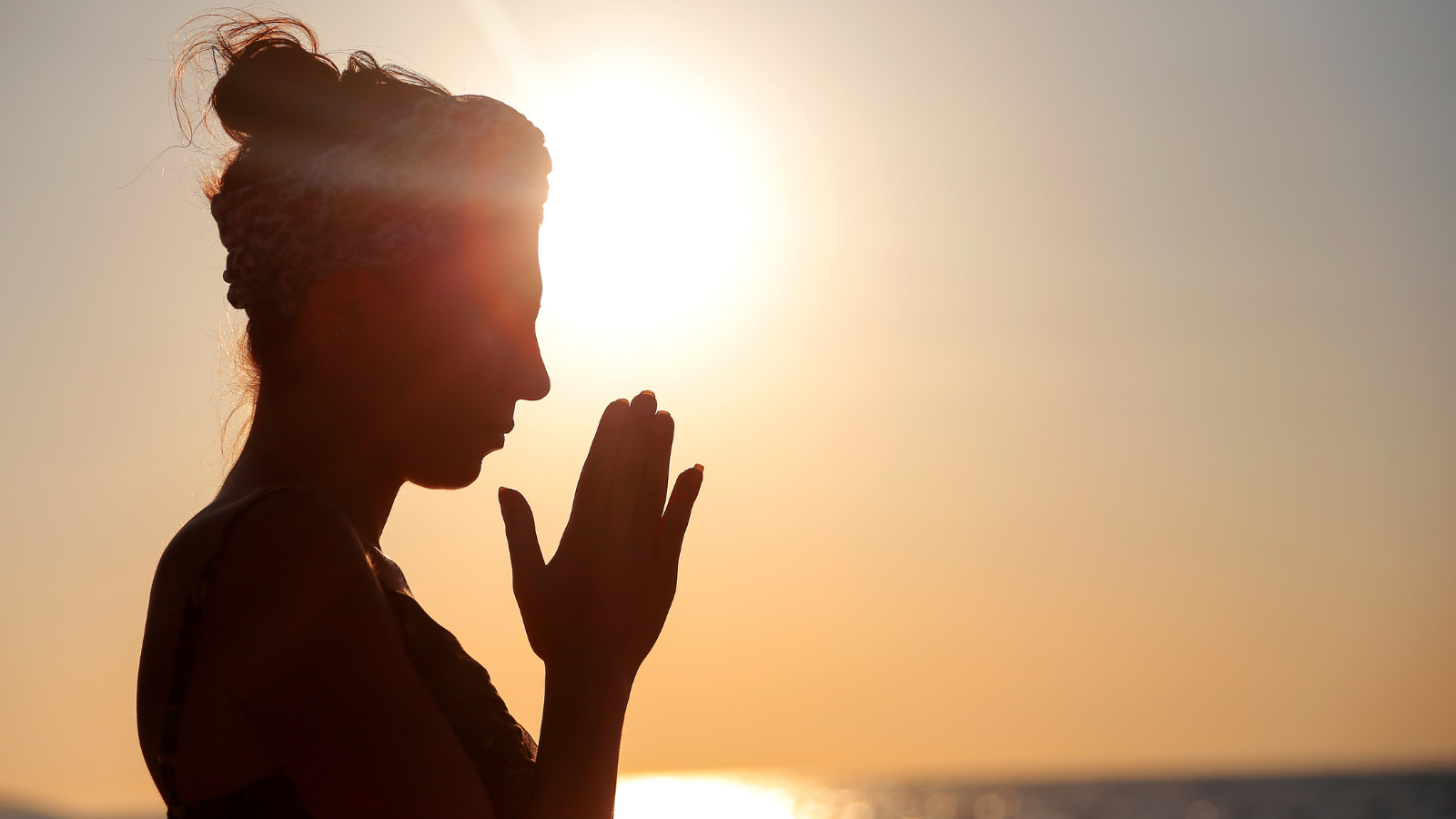 A woman standing on the beach at sunset with her hands in Anjali Mudra (Prayer Position), a gesture of self-healing.