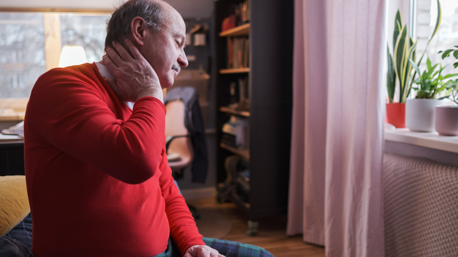 A senior man sitting in discomfort by a window in his home with one hand rubbing his neck. 